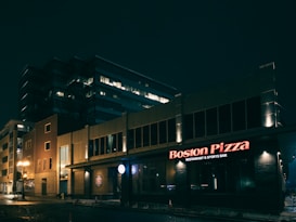 A nighttime urban scene featuring a well-lit restaurant and sports bar with the name 'Boston Pizza' displayed prominently in red neon lights. The building appears modern, with a large office structure in the background. Streetlights cast a warm glow on the wet pavement, suggesting recent rain.