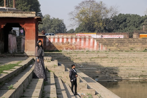 A man and a woman dressed in formal attire stand on stone steps next to a water body, possibly a stepwell or ghat. The woman is in a long, dark dress and the man is wearing a black suit. In the background, there is an old building with red and beige walls and signs written in Hindi. Trees and a clear sky can be seen further back.