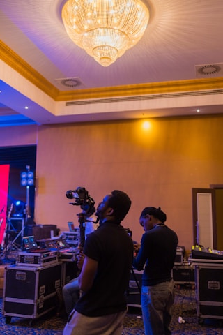 Two people are using professional video equipment in a venue with a large chandelier and various technical equipment around. The setting suggests preparation or coverage for an event.