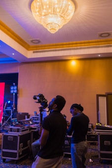 Two people are using professional video equipment in a venue with a large chandelier and various technical equipment around. The setting suggests preparation or coverage for an event.