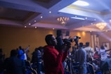 A person in a red hoodie is operating a video camera on a tripod in a dimly lit event space filled with people. The room is decorated with chandeliers and ambient lighting, creating a lively atmosphere as attendees engage with the event.