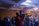 A person in a red hoodie is operating a video camera on a tripod in a dimly lit event space filled with people. The room is decorated with chandeliers and ambient lighting, creating a lively atmosphere as attendees engage with the event.