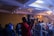 A person in a red hoodie is operating a video camera on a tripod in a dimly lit event space filled with people. The room is decorated with chandeliers and ambient lighting, creating a lively atmosphere as attendees engage with the event.