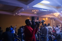 A person in a red hoodie is operating a video camera on a tripod in a dimly lit event space filled with people. The room is decorated with chandeliers and ambient lighting, creating a lively atmosphere as attendees engage with the event.