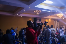A person in a red hoodie is operating a video camera on a tripod in a dimly lit event space filled with people. The room is decorated with chandeliers and ambient lighting, creating a lively atmosphere as attendees engage with the event.