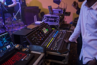 A person in a white shirt is controlling an audio mixing console in a dimly lit environment. Various electronic devices and displays with vibrant lights are present, indicating a live event or studio setting.