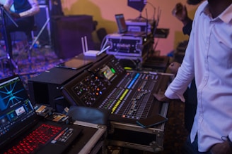 A person in a white shirt is controlling an audio mixing console in a dimly lit environment. Various electronic devices and displays with vibrant lights are present, indicating a live event or studio setting.
