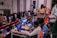 A group of people is gathered in an auditorium, with some seated and others standing. A person is operating a media setup with multiple screens and laptops on a desk, showing various video feeds. Camera equipment is set up nearby, indicating the event might be recorded or streamed. The environment appears lively and focused.