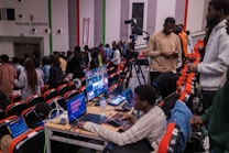 A group of people is gathered in an auditorium, with some seated and others standing. A person is operating a media setup with multiple screens and laptops on a desk, showing various video feeds. Camera equipment is set up nearby, indicating the event might be recorded or streamed. The environment appears lively and focused.