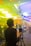 A person is using a camera on a tripod in a brightly lit indoor setting. The environment appears to be a conference or event with people sitting or standing in the background, which is out of focus. The lights cast colorful hues of yellow and blue across the room.