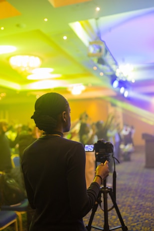 A person is using a camera on a tripod in a brightly lit indoor setting. The environment appears to be a conference or event with people sitting or standing in the background, which is out of focus. The lights cast colorful hues of yellow and blue across the room.