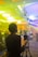 A person is using a camera on a tripod in a brightly lit indoor setting. The environment appears to be a conference or event with people sitting or standing in the background, which is out of focus. The lights cast colorful hues of yellow and blue across the room.