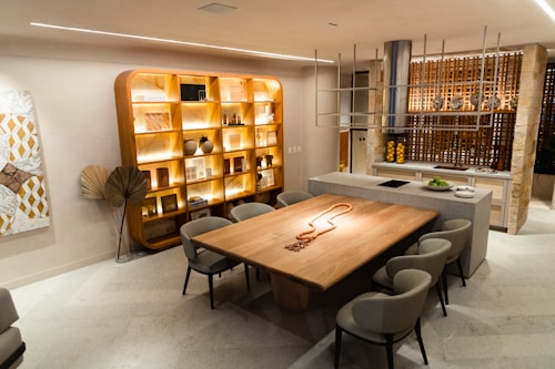 A modern dining area with a large wooden table surrounded by six upholstered chairs. The room features a backlit wooden shelving unit displaying various decorative objects, such as vases and books. A contemporary kitchen island with fruit and a pendant chandelier adds a stylish touch. Warm lighting creates a cozy ambiance, complemented by artwork on the wall.