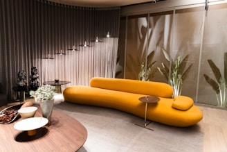 A modern interior featuring a mustard yellow, elongated sofa with a unique, abstract design. It is positioned against a backdrop of metallic curtains and a wall adorned with large potted plants. The room has a minimalist aesthetic, marked by circular hanging lights above and a round wooden coffee table in the foreground, adorned with decorative objects and a silver vase with flowers.