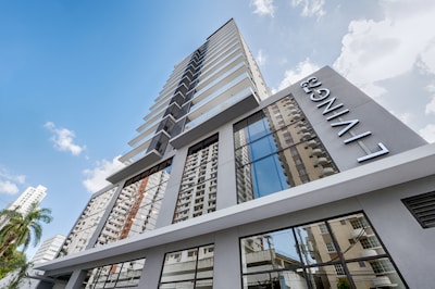 A modern high-rise building with a sleek design featuring large glass windows reflecting other nearby buildings. The name 'LIVING' is prominently displayed on the side of the building. The sky is mostly clear with a few scattered clouds, and some palm trees are visible near the base.