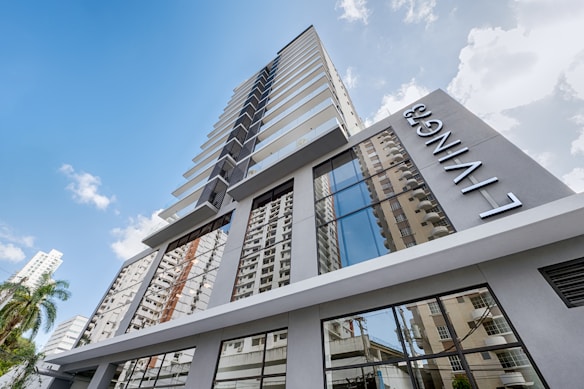 A modern high-rise building with a sleek design featuring large glass windows reflecting other nearby buildings. The name 'LIVING' is prominently displayed on the side of the building. The sky is mostly clear with a few scattered clouds, and some palm trees are visible near the base.