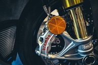 A close-up view of a motorcycle's front wheel assembly, featuring a silver brake caliper labeled 'Brembo' and a prominent golden suspension fork. An orange reflector and Ohlins branding are also visible.