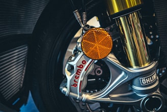 A close-up view of a motorcycle's front wheel assembly, featuring a silver brake caliper labeled 'Brembo' and a prominent golden suspension fork. An orange reflector and Ohlins branding are also visible.