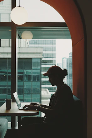 A person working on a laptop in a cozy home office with a city skyline visible through the window.