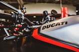 Close-up of a motorcycle with the brand name Ducati visible on the metallic tank, highlighting the sleek design and bold red accent. The handlebars and parts of the garage setting are also visible in the background.