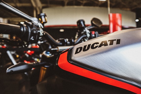 Close-up of a motorcycle with the brand name Ducati visible on the metallic tank, highlighting the sleek design and bold red accent. The handlebars and parts of the garage setting are also visible in the background.