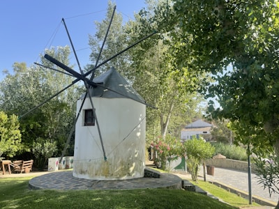 A traditional windmill surrounded by lush greenery, with various trees and bushes. The windmill has a white, cylindrical body with a conical roof and several blades projecting from its top. Nearby, a wooden bench and several decorative elements enhance the quaint, rustic setting. A paved pathway runs alongside the windmill, leading toward a building in the background.