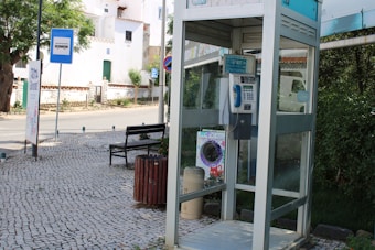 A street scene featuring a public telephone booth with glass walls and a keypad inside. The booth is located on a cobblestone sidewalk with a trash can nearby. In the background, there is a bench, a bus stop sign, a road sign, and a white building with green doors and windows. Trees and greenery are present, adding a touch of nature to the urban environment.