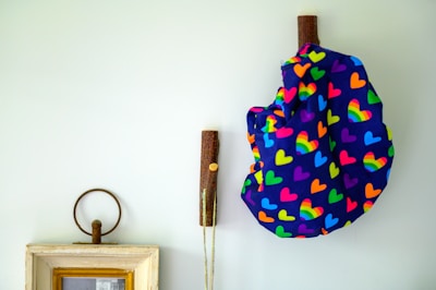 A wall features decorative elements including a patterned cloth or bag adorned with colorful hearts and rainbow designs, hanging on rustic wooden pegs. A framed object below shares the space, adding a vintage touch with its minimalistic frame and faded photograph.