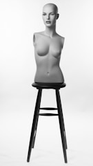 A female mannequin torso with a bald head and realistic facial features is positioned upright on a tall black stool. It is lit against a simple white background, highlighting its form and features.