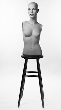 A female mannequin torso with a bald head and realistic facial features is positioned upright on a tall black stool. It is lit against a simple white background, highlighting its form and features.