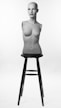 A female mannequin torso with a bald head and realistic facial features is positioned upright on a tall black stool. It is lit against a simple white background, highlighting its form and features.