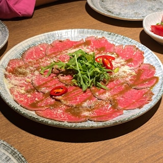 A plate of thinly sliced raw beef, garnished with arugula, sliced red chili peppers, and a sprinkle of cheese. The beef is arranged neatly in a circular pattern on a textured plate with some sauce drizzled on top.