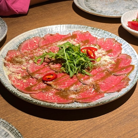 A plate of thinly sliced raw beef, garnished with arugula, sliced red chili peppers, and a sprinkle of cheese. The beef is arranged neatly in a circular pattern on a textured plate with some sauce drizzled on top.