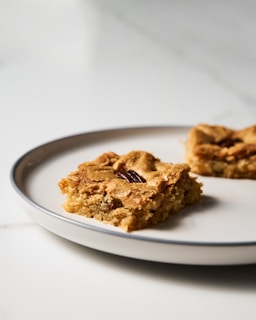 A square dessert with a golden brown crust sits on a round white plate. The baked item appears to have pieces of nuts embedded in it and has a crumbly texture.