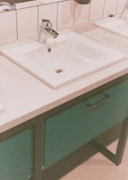 A modern bathroom sink with a chrome faucet set in a rectangular countertop with white tiles in the background. The basin is surrounded by a smooth counter surface, and below it is a cabinet with a teal color finish and a black handle.