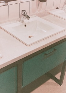 A modern bathroom sink with a chrome faucet set in a rectangular countertop with white tiles in the background. The basin is surrounded by a smooth counter surface, and below it is a cabinet with a teal color finish and a black handle.