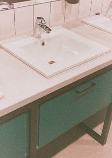 A modern bathroom sink with a chrome faucet set in a rectangular countertop with white tiles in the background. The basin is surrounded by a smooth counter surface, and below it is a cabinet with a teal color finish and a black handle.