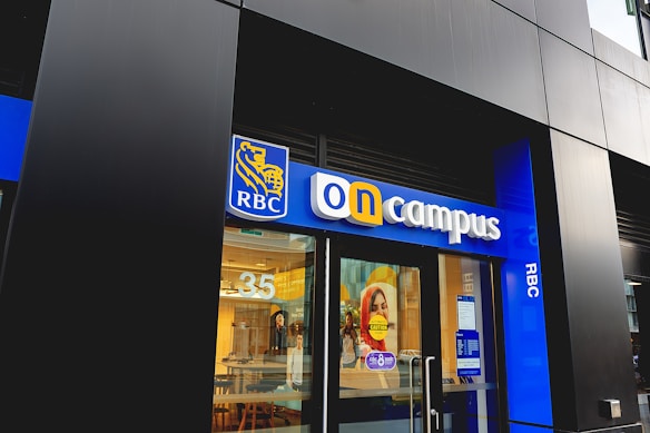 A storefront with a modern design, featuring a vibrant blue sign with the letters 'RBC' on it. Below 'RBC', large letters spell out 'on campus', with a glass door entrance. Inside, the interior appears well-lit with a warm, inviting atmosphere. The number '35' is visible on the glass, and signage with information is displayed.