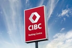 A large outdoor sign with a red background displaying the logo and text of a banking center, set against a partly cloudy blue sky.