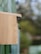 A wooden birdhouse is mounted outdoors with a bee or similar insect flying near the entrance. The background is blurred, suggesting a natural, outdoor setting with greenery.