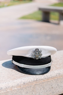 A white military-style cap with a black brim and a metallic emblem displaying an eagle is placed on a stone surface. The background is blurred, showing hints of a park with grass and benches.