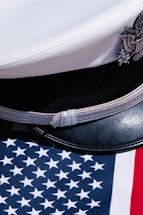 A white naval officer's cap with an emblem rests on an American flag that features blue with white stars and red and white stripes.