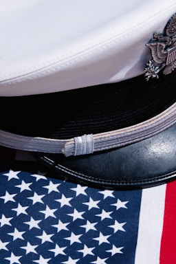 A white naval officer's cap with an emblem rests on an American flag that features blue with white stars and red and white stripes.
