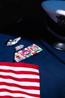 A detailed view of a military uniform displaying various colorful ribbons and medals. A naval officer's hat, with a black visor and white cover featuring an emblem, rests nearby. A striped pattern in red and white, resembling part of a flag, is partially visible.