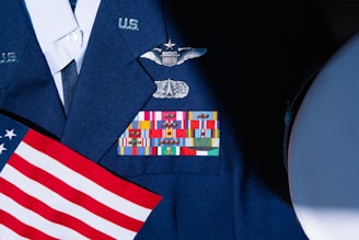 A military uniform with various colorful ribbons and medals displayed on it, accompanied by a small American flag. The uniform is dark blue with metallic badges indicating rank and service.