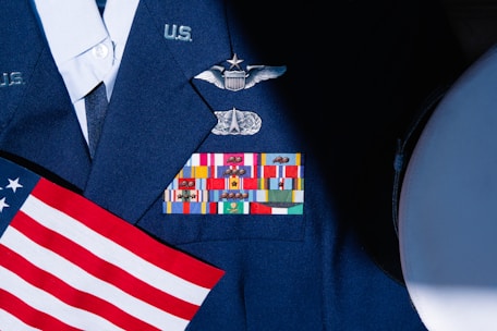 A military uniform with various colorful ribbons and medals displayed on it, accompanied by a small American flag. The uniform is dark blue with metallic badges indicating rank and service.