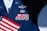 A military uniform with various colorful ribbons and medals displayed on it, accompanied by a small American flag. The uniform is dark blue with metallic badges indicating rank and service.