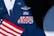 A military uniform with various colorful ribbons and medals displayed on it, accompanied by a small American flag. The uniform is dark blue with metallic badges indicating rank and service.