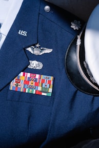 A formal military uniform featuring a collection of service ribbons, pins, and insignia on a navy blue coat. Visible is a white peaked cap with a black visor beside it. The uniform displays symbols of rank and recognition.
