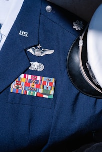 A formal military uniform featuring a collection of service ribbons, pins, and insignia on a navy blue coat. Visible is a white peaked cap with a black visor beside it. The uniform displays symbols of rank and recognition.